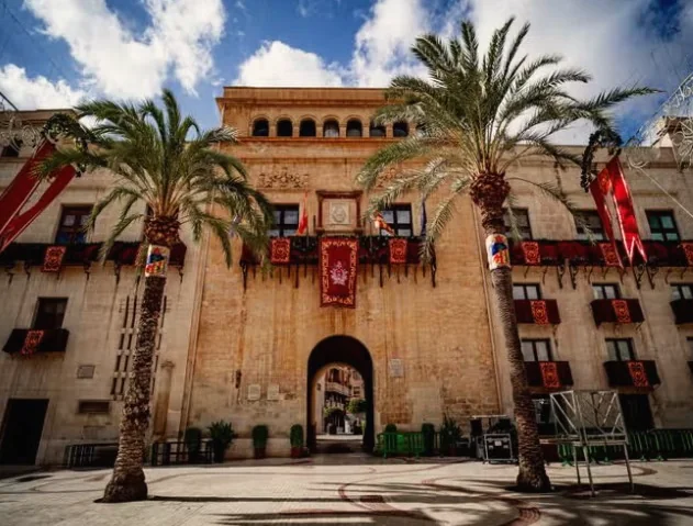 A sandstone civic building with palms and red banners in Elche’s old town